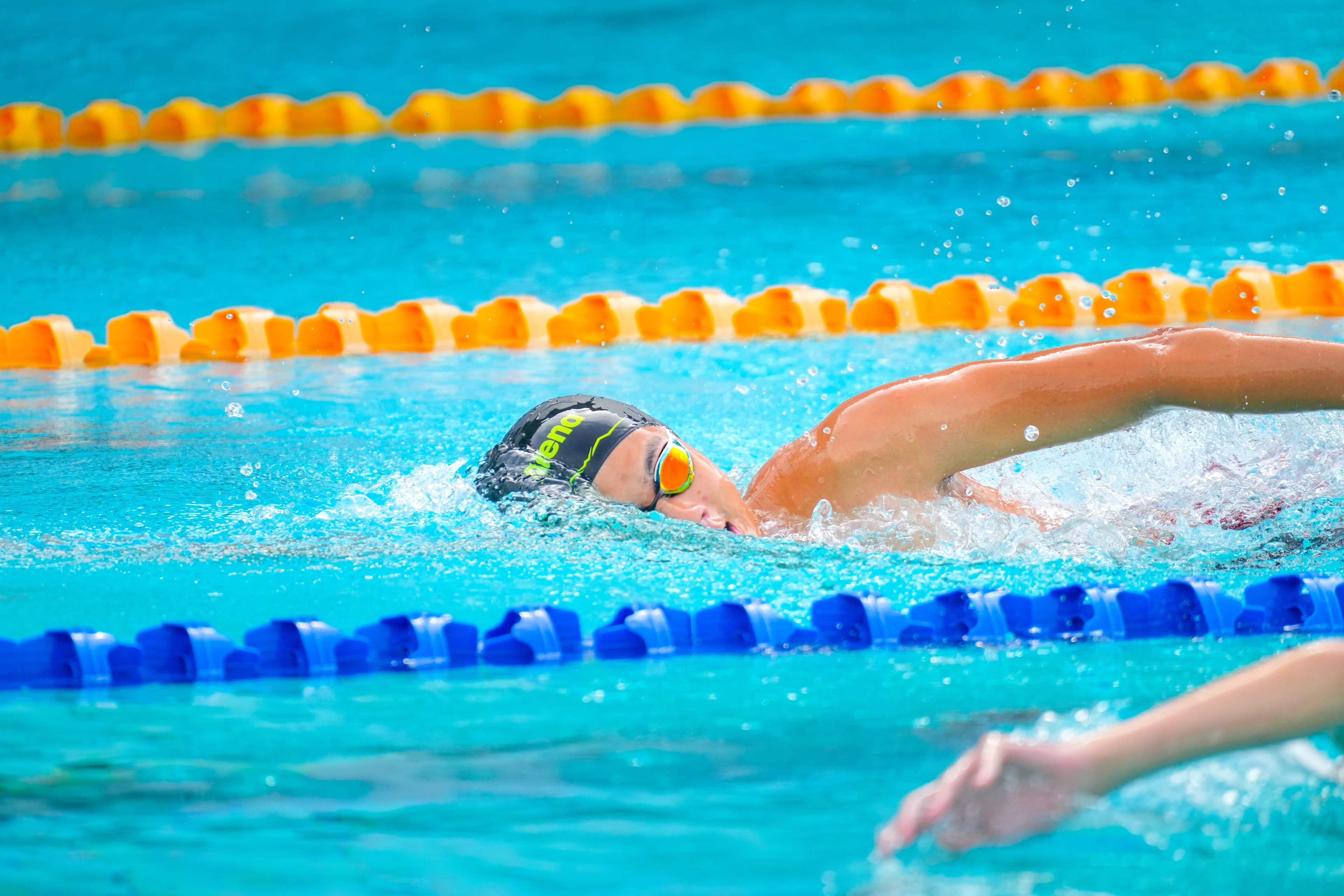 A swimmer mid-freestyle stroke, head turned for breath, orange and blue lane ropes visible across the pool.