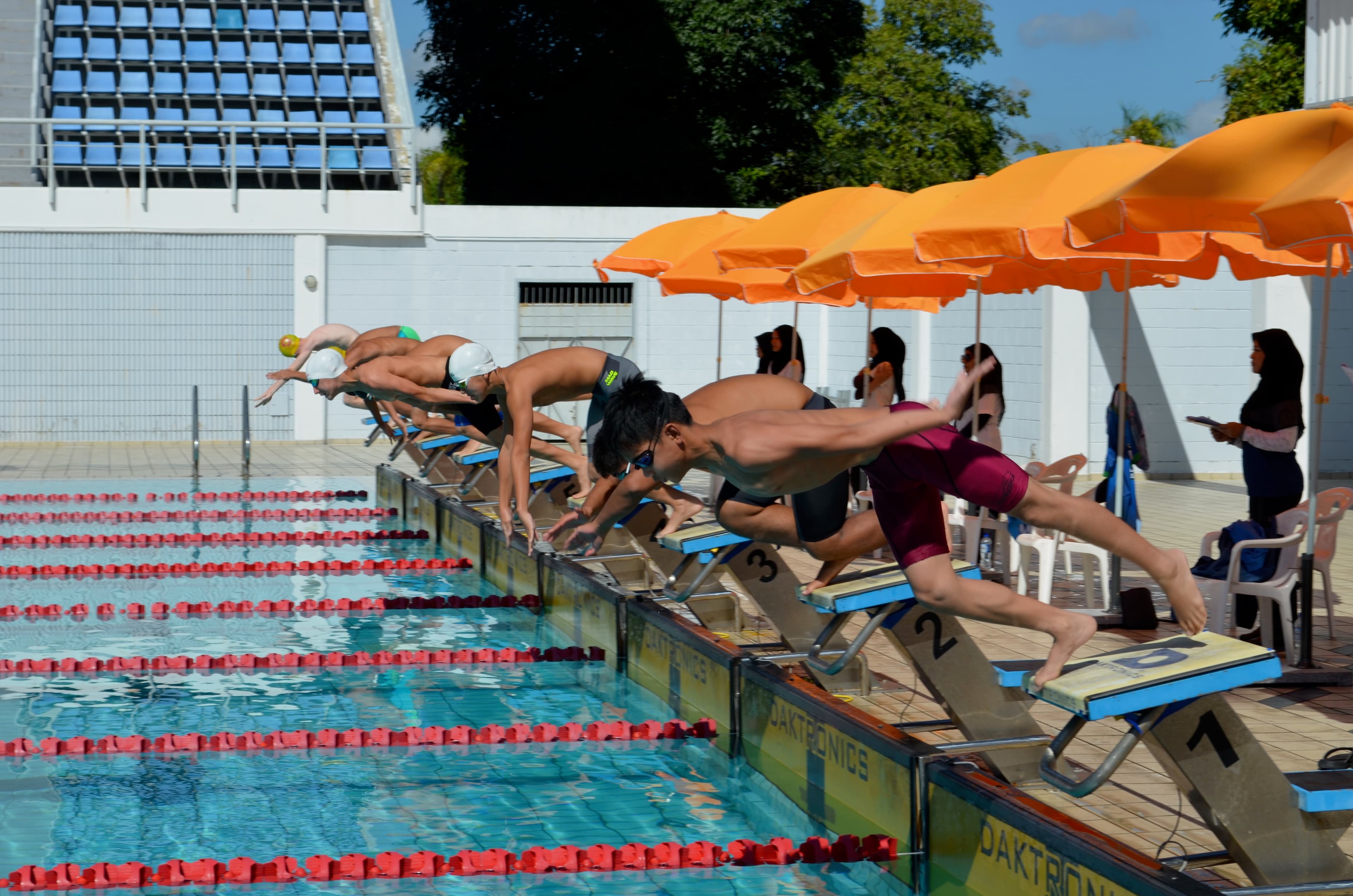 Five swimmers diving from starting blocks at the Hassanal Bolkiah National Swimming Complex, with race officials seated under an orange umbrella to the right.